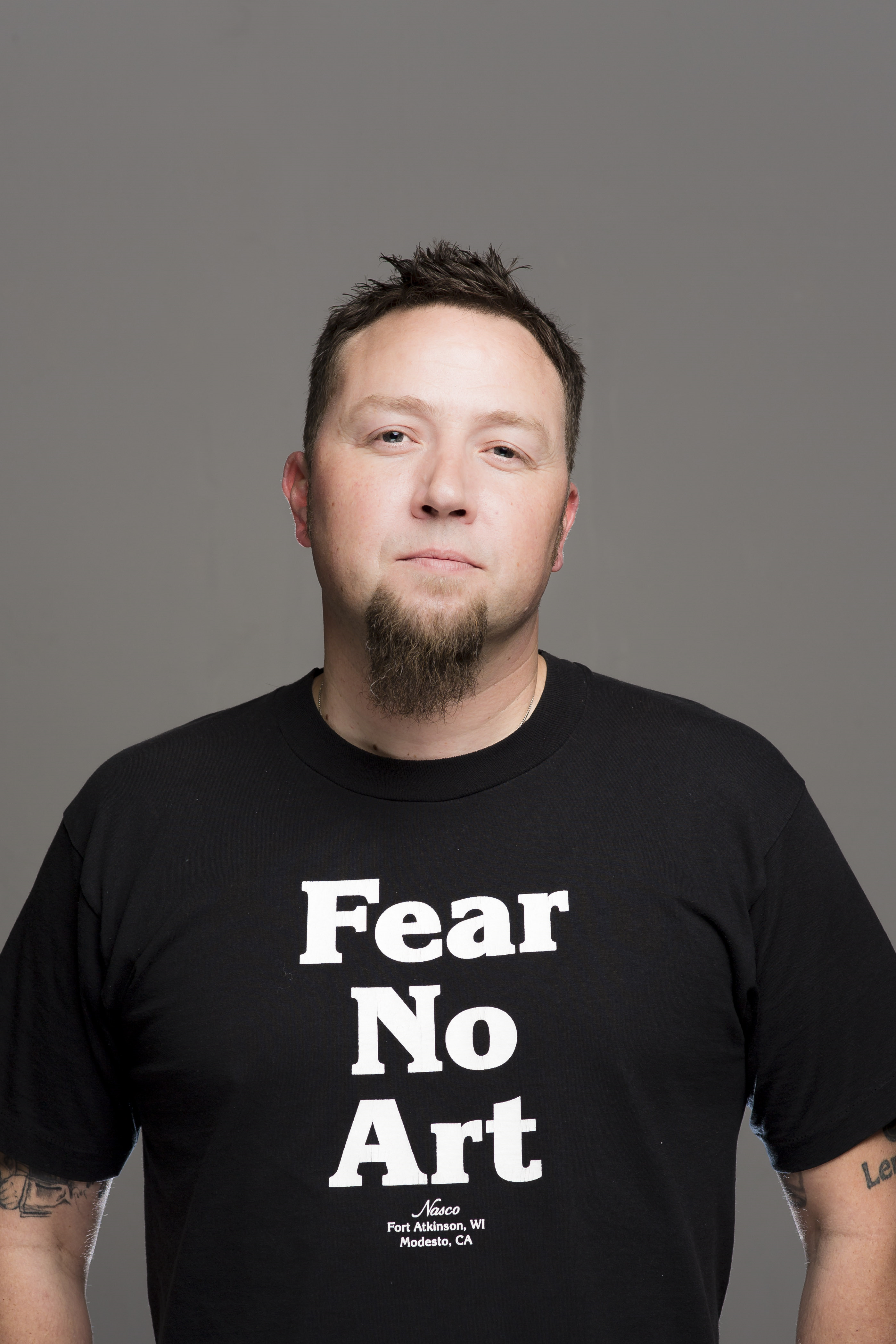 Image from the waist up of Ray McManus wearing a black t-shirt with the words "Fear No Art" across the front in white letters.