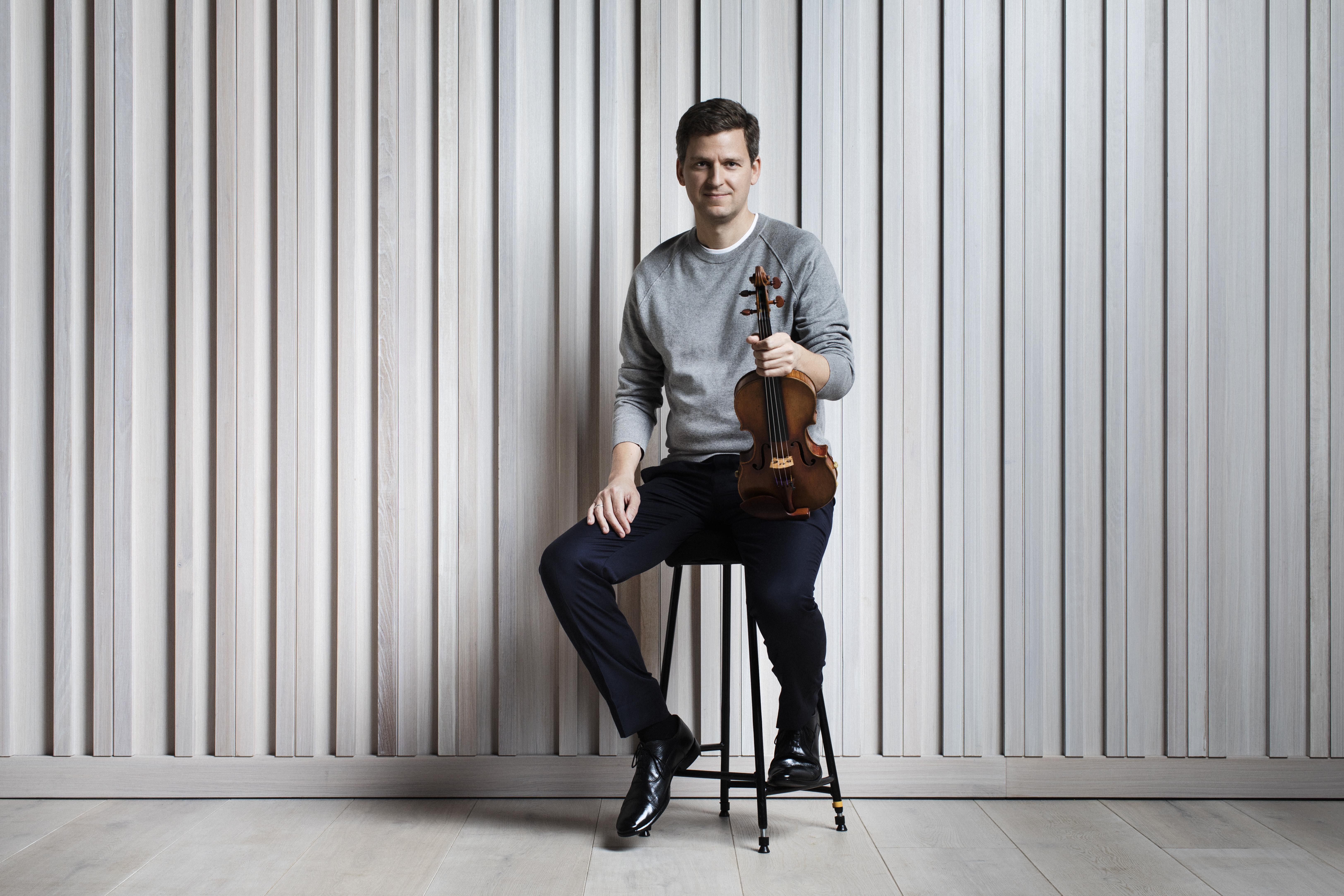 A man in a gray sweater holding a violin and sitting on a stool against a beige wall