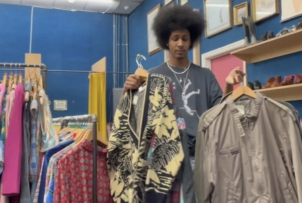 a man standing in a vintage clothing shop holding up a sweater and a jacket