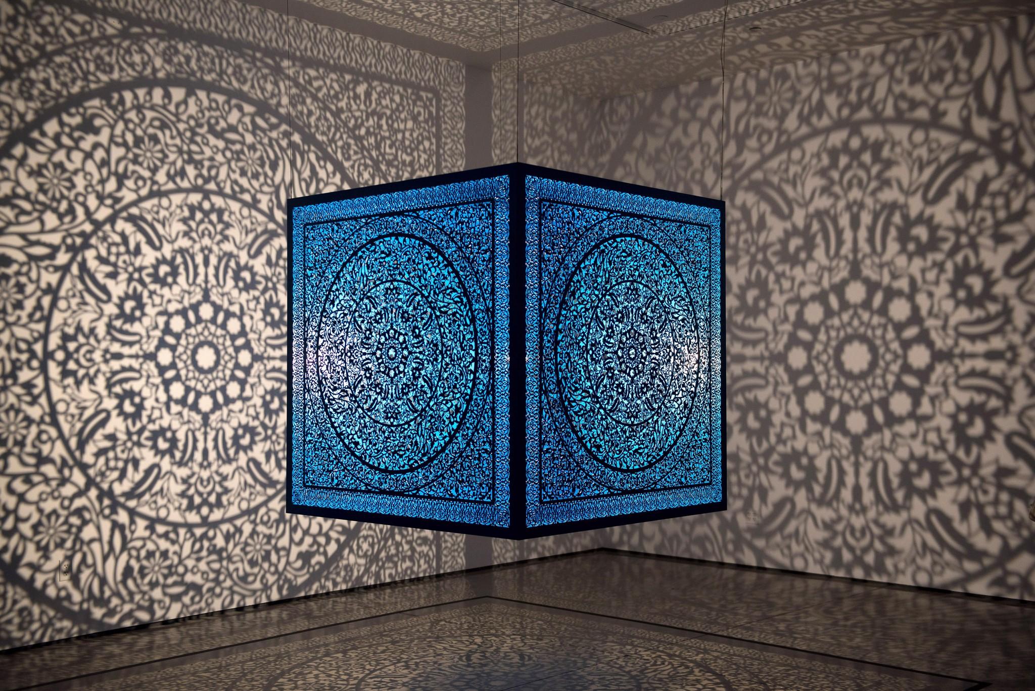 A blue steel cube with intricate floral-inspired cutouts suspended from the ceiling and casting patterned light onto the gray walls of a room.