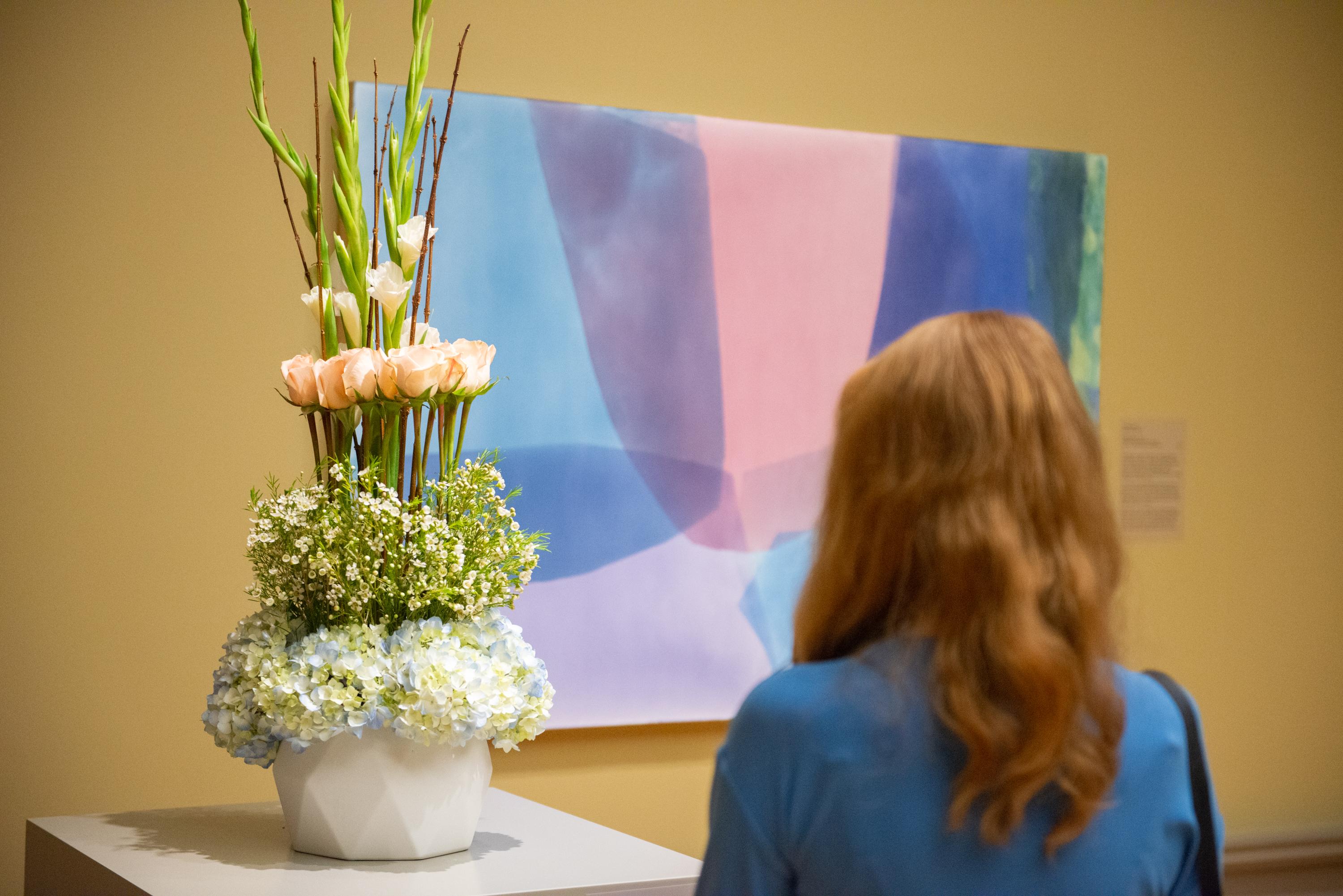 Woman in the CMA galleries with her back to camera facing a painting in pastel colors and a floral arrangement interpreting the painting.