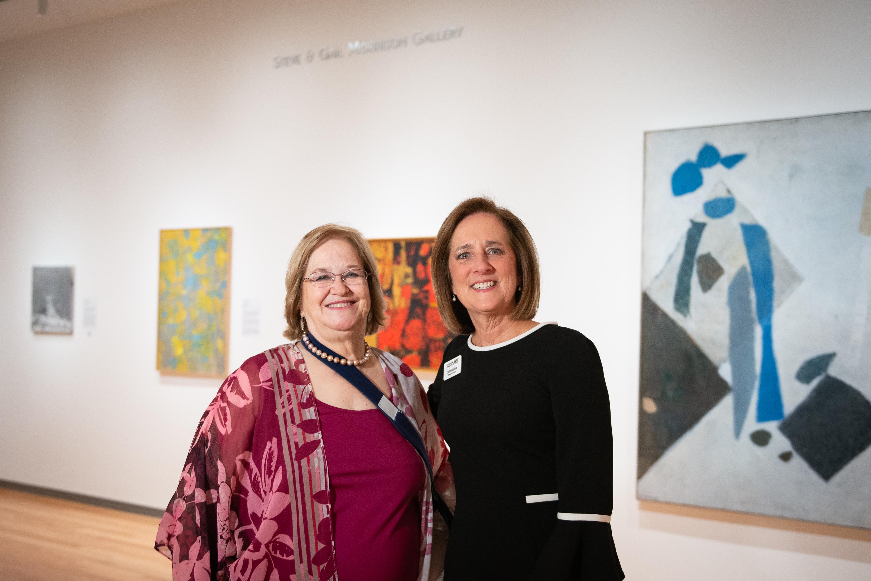 Two women, Dr. Gail Morrison and Della Watkins, smiling in front of a gallery wall hung with contemporary abstract paintings.
