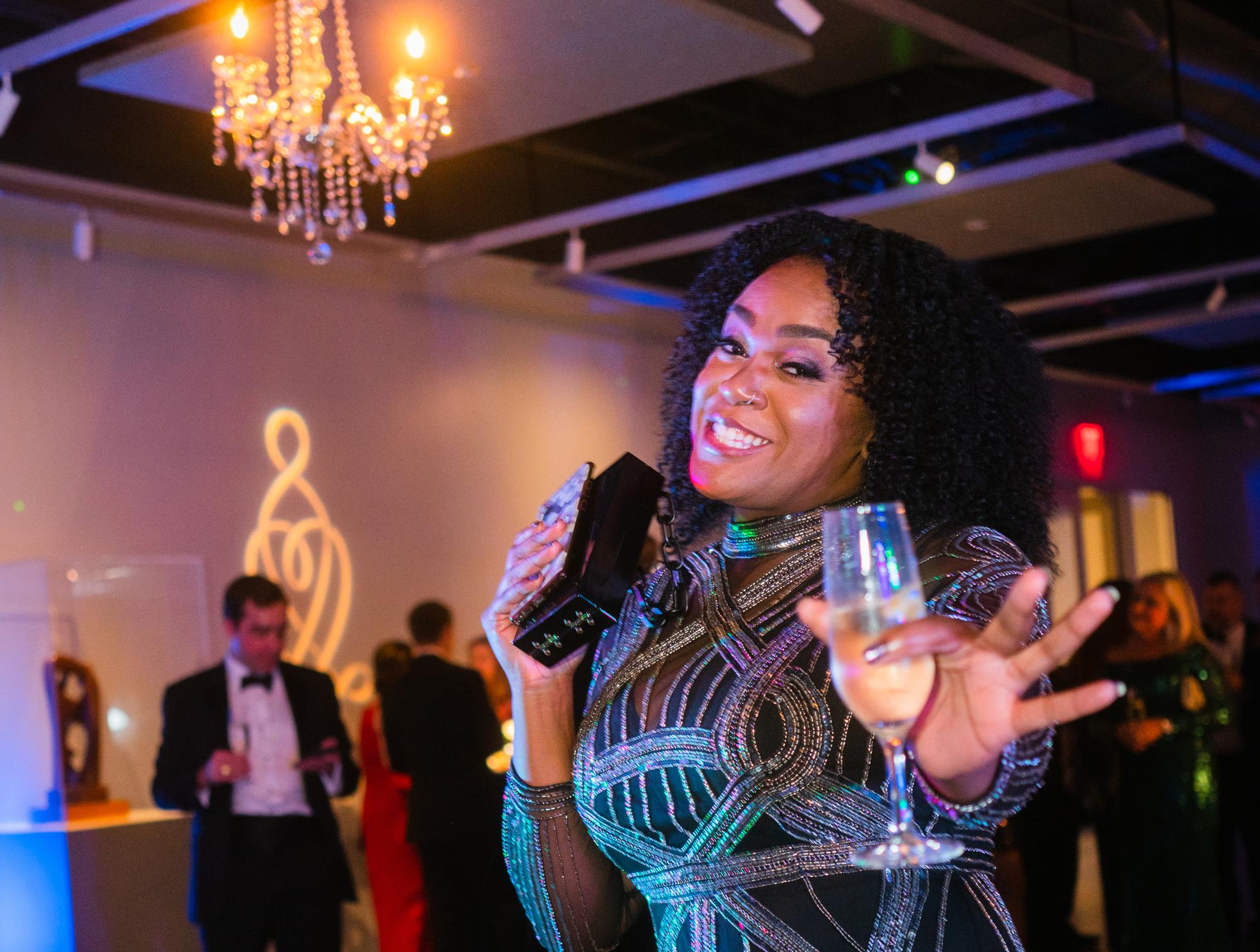 Image of woman at last year's gala in a sparkly dress smiling and gesturing to camera with a glass of champagne