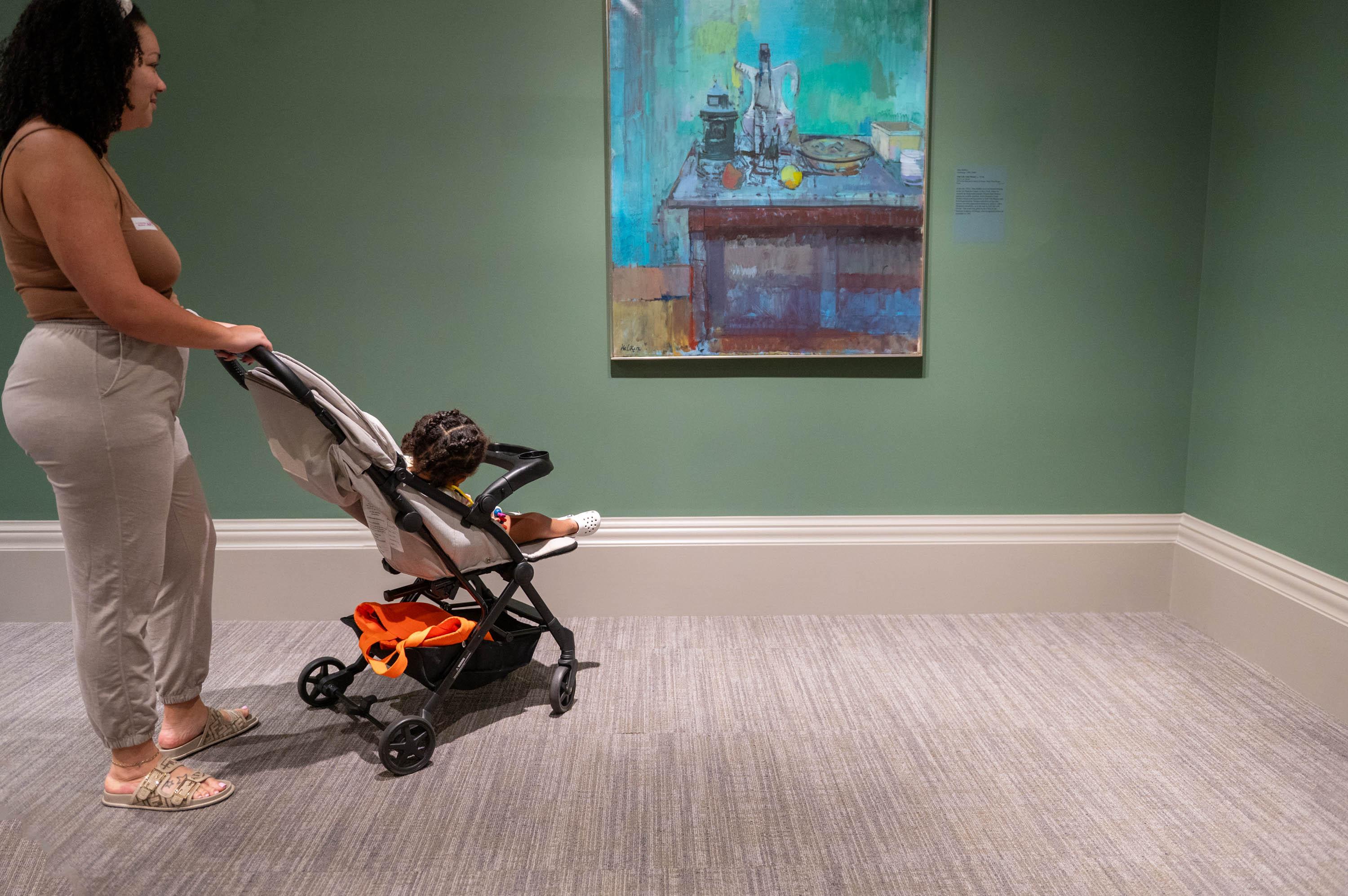 Woman of color with toddler in stroller looking at colorful art painting 