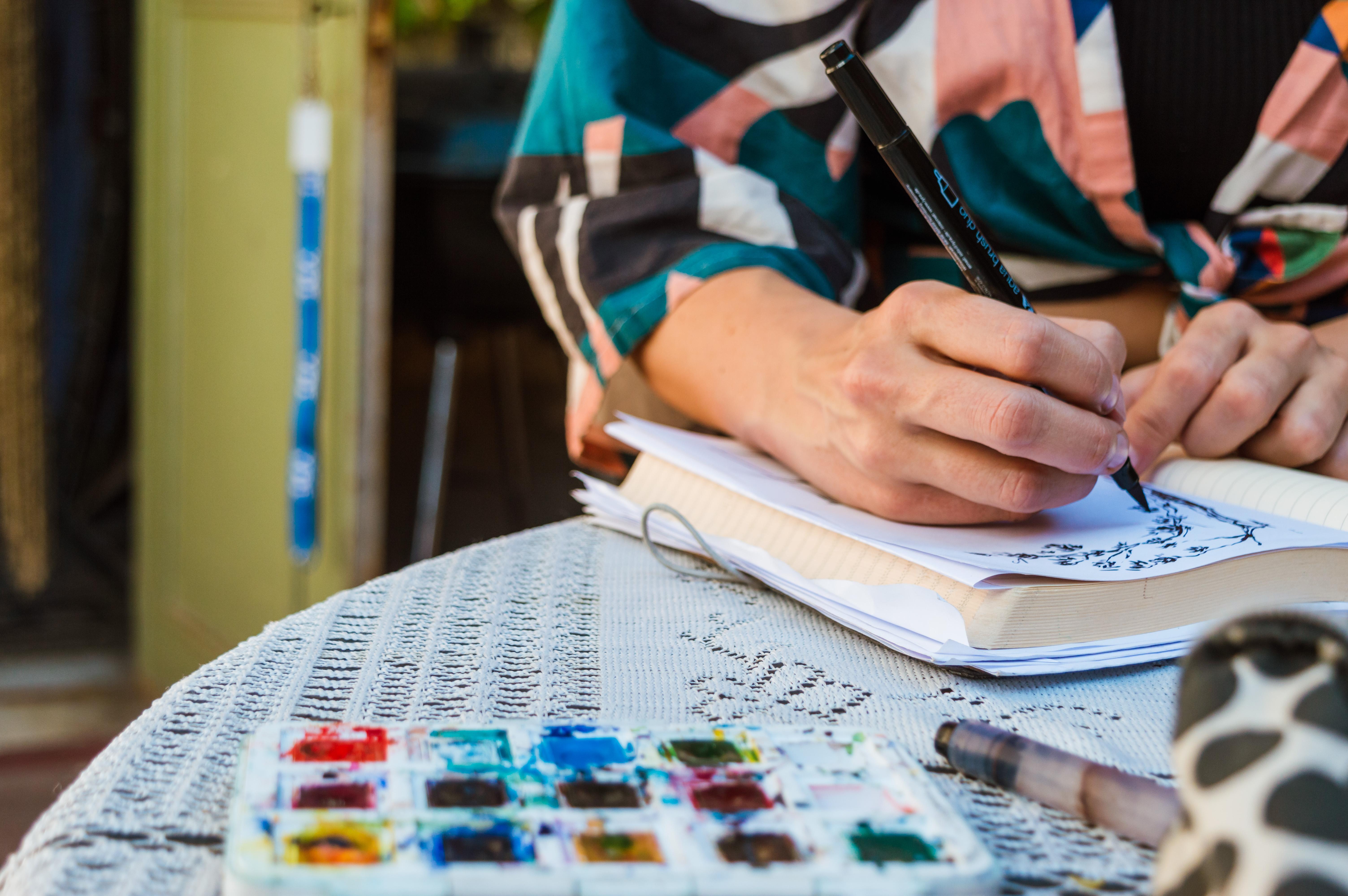 A woman sketching in her journal with paint supplies at hand