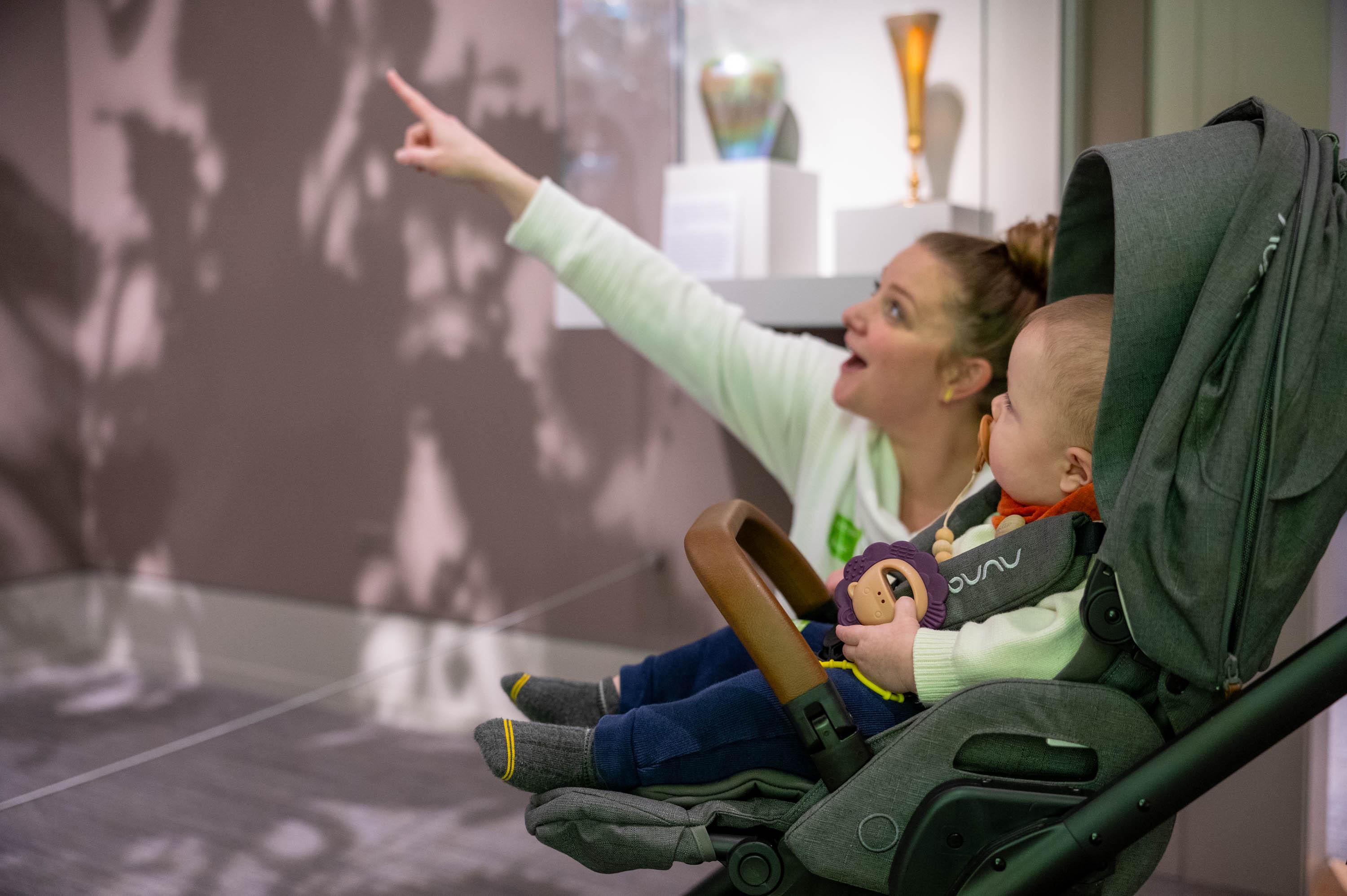 a caregiver lowered to eye-level with a child in a stroller points out nearby art to the child