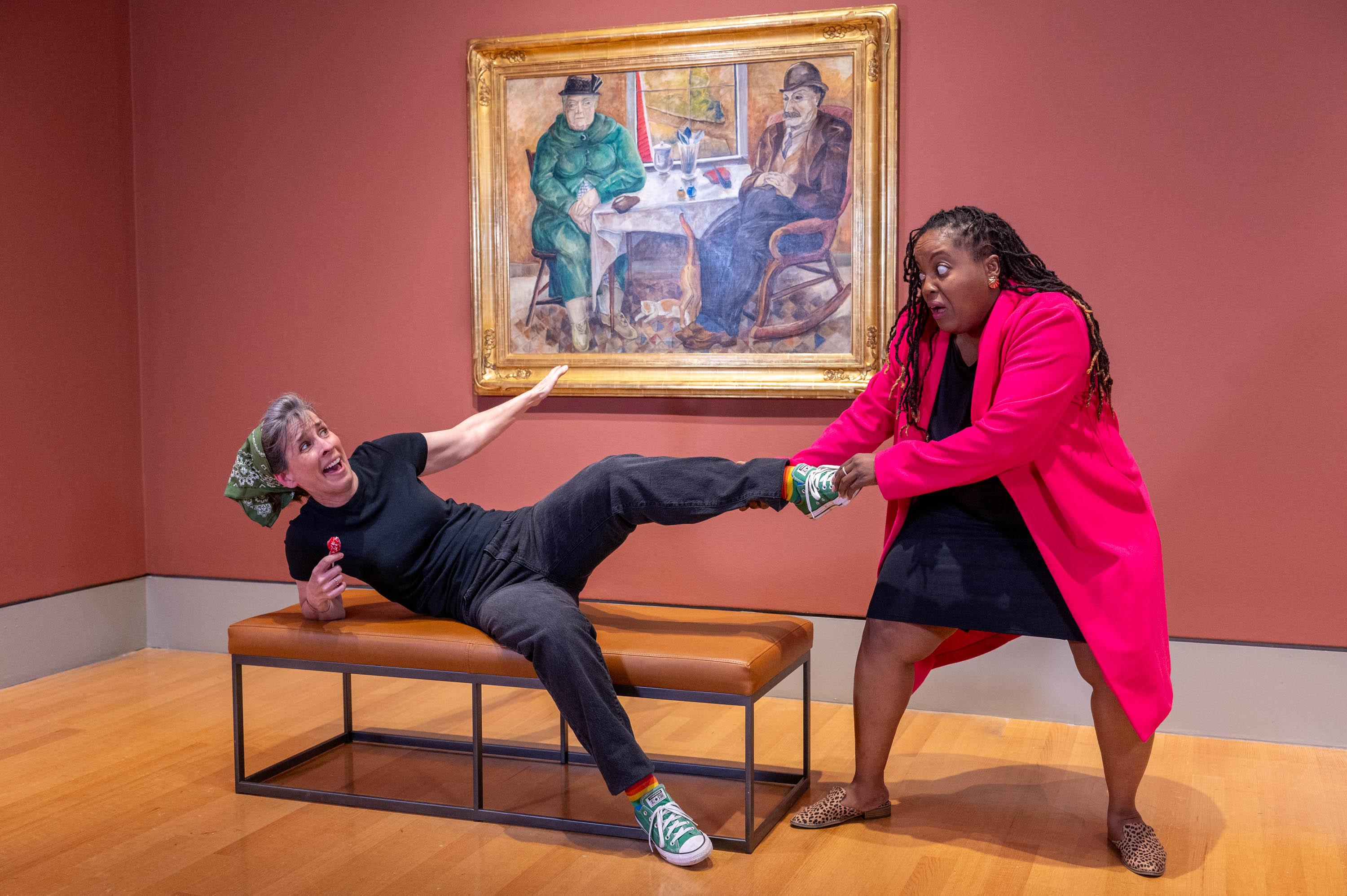Two actors are in the galleries. One lies on a bench while the other pulls her by her foot